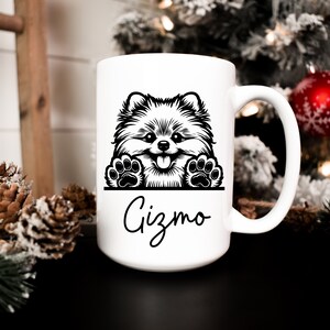 K&ouml;nnte beinhalten: Wei&szlig;e Keramiktasse mit einer Schwarzwei&szlig;illustration eines Zwergspitz-Hundes, der &uuml;ber den Rand schaut. Die Tasse hat einen schwarzen Henkel und den Namen "Gizmo" in Kursivschrift unter dem Hund.