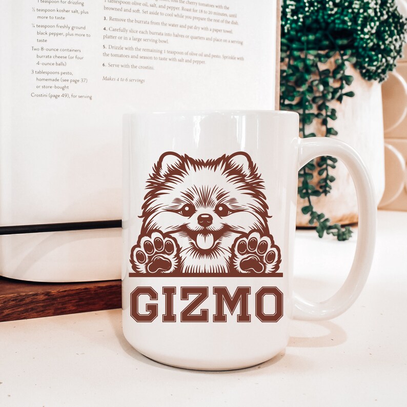 K&ouml;nnte beinhalten: Eine wei&szlig;e Keramiktasse mit einer braunen Illustration eines Zwergspitz-Hundes, der &uuml;ber den Rand schaut. Der Hund hat eine braune Nase, Zunge und Pfoten. Der Text "GIZMO" ist in braunen Buchstaben unter dem Hund gedruckt.