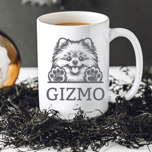 K&ouml;nnte beinhalten: Wei&szlig;e Keramiktasse mit einer Schwarzwei&szlig;illustration eines Zwergspitz-Hundes, der &uuml;ber den Rand schaut. Der Text "Gizmo" ist unter dem Hund aufgedruckt.
