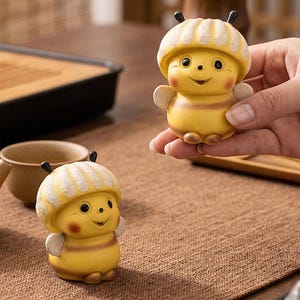 May include: Two yellow and cream-colored bee figurines with smiling faces and small wings. One figurine is held in a hand, while the other sits on a brown surface. A teapot and teacup are also visible, suggesting a tea-themed item.