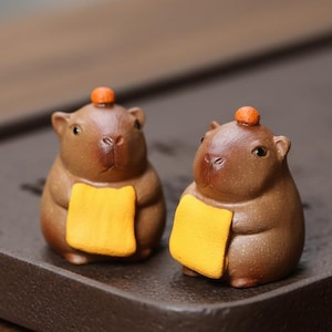 May include: Two brown ceramic capybara figurines with orange accents and yellow bibs. Each figurine has a small orange sphere on its head. The capybaras are on a dark brown tray.