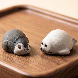 May include: Two ceramic figurines: a penguin and a seal. The penguin is dark gray with a white belly and face, and the seal is white with dark gray details. Both are on a wooden tray.