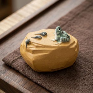 May include: A ceramic frog figurine with a light green and white pattern sits on a tan-colored rock, alongside three small tadpoles. The frog is detailed with visible eyes and legs. This decorative piece is likely a home decor item.