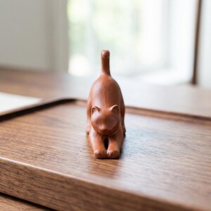 Puede incluir: Una figura de gato marr&oacute;n en posici&oacute;n de estiramiento. El gato est&aacute; hecho de un material s&oacute;lido y tiene un acabado liso. La figura est&aacute; sobre una bandeja de madera con un acabado marr&oacute;n oscuro. La cola del gato est&aacute; levantada.