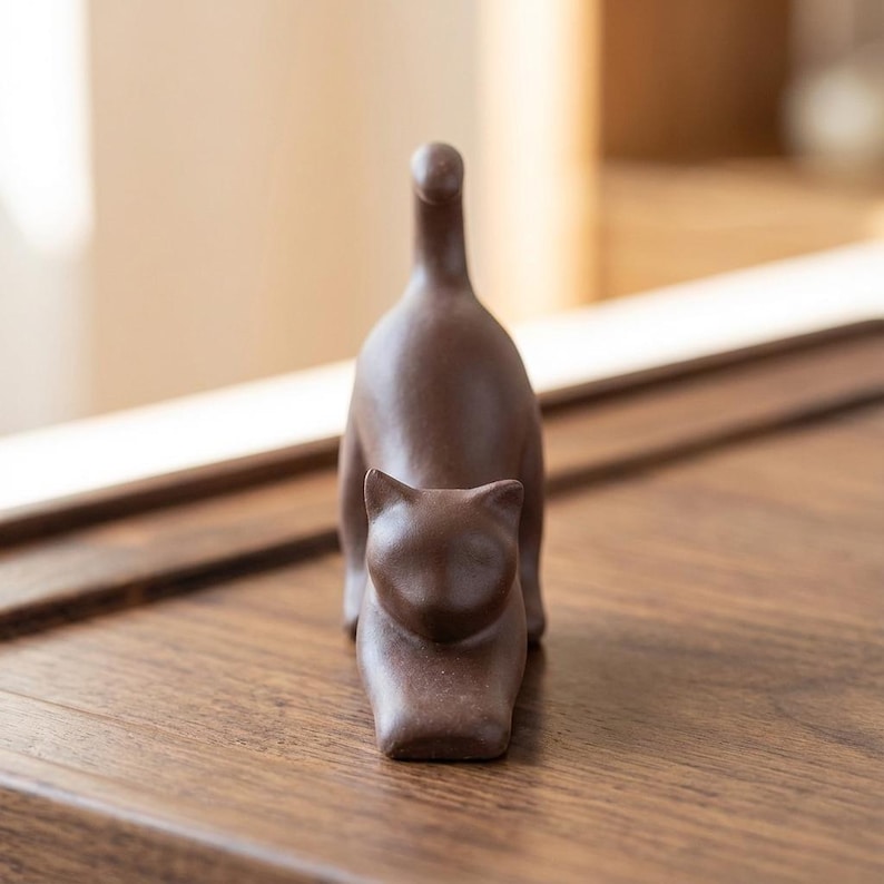 Puede incluir: Una figura de gato de cer&aacute;mica marr&oacute;n en una pose juguetona, con la cola levantada. El gato est&aacute; representado en un estilo minimalista, con rasgos suaves y redondeados. La figura est&aacute; sobre una superficie de madera.