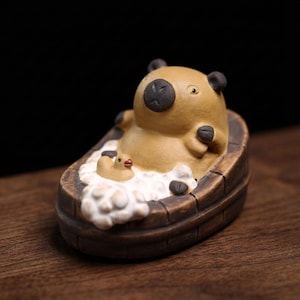 May include: A ceramic figurine of a capybara relaxing in a wooden bathtub filled with white bubbles. A small yellow duck figurine floats in the bubbles. The capybara has black ears and a black nose.