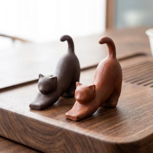 Puede incluir: Dos figuras de gato de cer&aacute;mica en pose de estiramiento. Una es de color marr&oacute;n oscuro y la otra es de color marr&oacute;n rojizo. Las figuras de gato est&aacute;n sobre una superficie de madera. Las figuras de gato son un objeto decorativo.