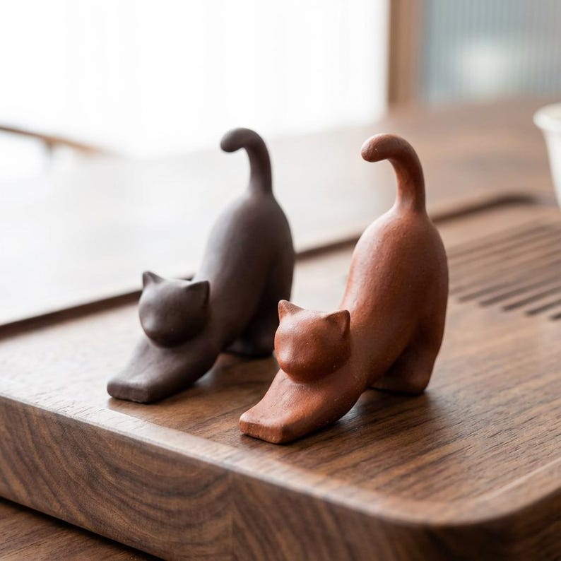 Puede incluir: Dos figuras de gato de cer&aacute;mica en pose de estiramiento. Una es de color marr&oacute;n oscuro y la otra es de color marr&oacute;n rojizo. Las figuras de gato est&aacute;n sobre una superficie de madera. Las figuras de gato son un objeto decorativo.