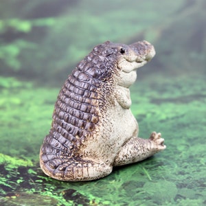 May include: A ceramic figurine of a crocodile with a textured, brown and gray body. The crocodile is sitting with its legs tucked under its body and its head tilted to the side.