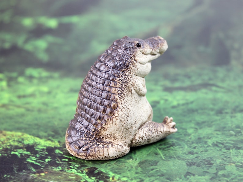 May include: A ceramic figurine of a crocodile with a textured, brown and gray body. The crocodile is sitting with its legs tucked under its body and its head tilted to the side.