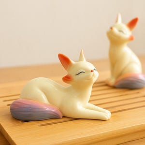 Könnte beinhalten: Zwei cremefarbene Keramik-Katzenfiguren mit orangefarbenen Ohren und einem Regenbogen-Schwanz. Eine Katze liegt, die andere sitzt. Sie befinden sich auf einer hellbraunen Holzoberfläche.