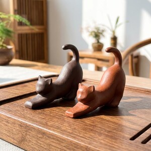 Puede incluir: Dos figuras de gato de cer&aacute;mica en pose de estiramiento. Una es de color marr&oacute;n oscuro y la otra marr&oacute;n rojizo. Se exhiben sobre una superficie de madera, mostrando su detallada artesan&iacute;a y dise&ntilde;o l&uacute;dico.