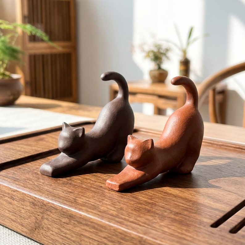 Puede incluir: Dos figuras de gato de cer&aacute;mica en pose de estiramiento. Una es de color marr&oacute;n oscuro y la otra marr&oacute;n rojizo. Se exhiben sobre una superficie de madera, mostrando su detallada artesan&iacute;a y dise&ntilde;o l&uacute;dico.