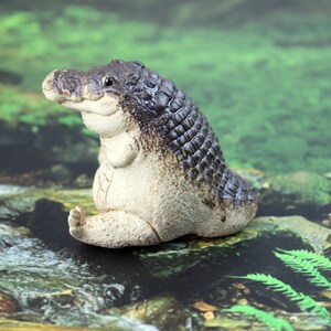 May include: A small, ceramic figurine of a crocodile sitting with its legs crossed. The crocodile is painted in shades of brown and white, with a textured surface that resembles scales. The figurine is sitting on a mossy green surface.