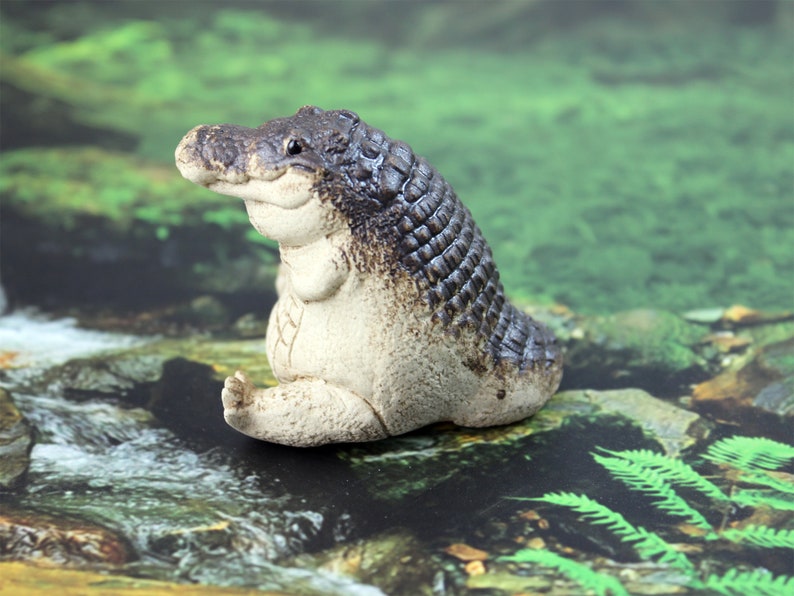 May include: A small, ceramic figurine of a crocodile sitting with its legs crossed. The crocodile is painted in shades of brown and white, with a textured surface that resembles scales. The figurine is sitting on a mossy green surface.