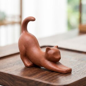 Puede incluir: Figura de gato de color terracota en pose de estiramiento. El gato est&aacute; arqueado, con las patas delanteras extendidas y la cola curvada hacia arriba. La figura est&aacute; sobre una bandeja de madera oscura, mostrando la forma suave y detallada del gato.