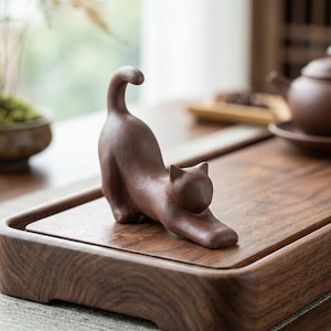 Puede incluir: Figura de gato de cer&aacute;mica marr&oacute;n en posici&oacute;n de estiramiento. El gato est&aacute; sobre una bandeja de madera, con una tetera y otros objetos en el fondo. La figura tiene un acabado liso y mate y es un objeto decorativo.