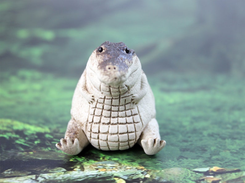 May include: A white ceramic alligator figurine with a textured, scaly body and black eyes. The alligator is sitting upright with its mouth closed and its front legs crossed.