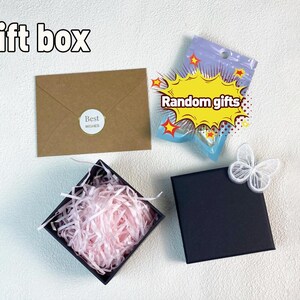 May include: A black gift box with pink tissue paper inside. A brown envelope with the text "Best Wishes" is in the background. A small plastic bag with the text "Random gifts" is also in the background.