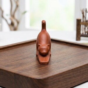 Puede incluir: Una peque&ntilde;a figura de gato marr&oacute;n rojizo en pose de estiramiento. El gato est&aacute; sobre una bandeja rectangular de madera. La bandeja tiene esquinas redondeadas y un acabado marr&oacute;n oscuro. El fondo est&aacute; borroso, con una ventana y algunos elementos decorativos visibles.