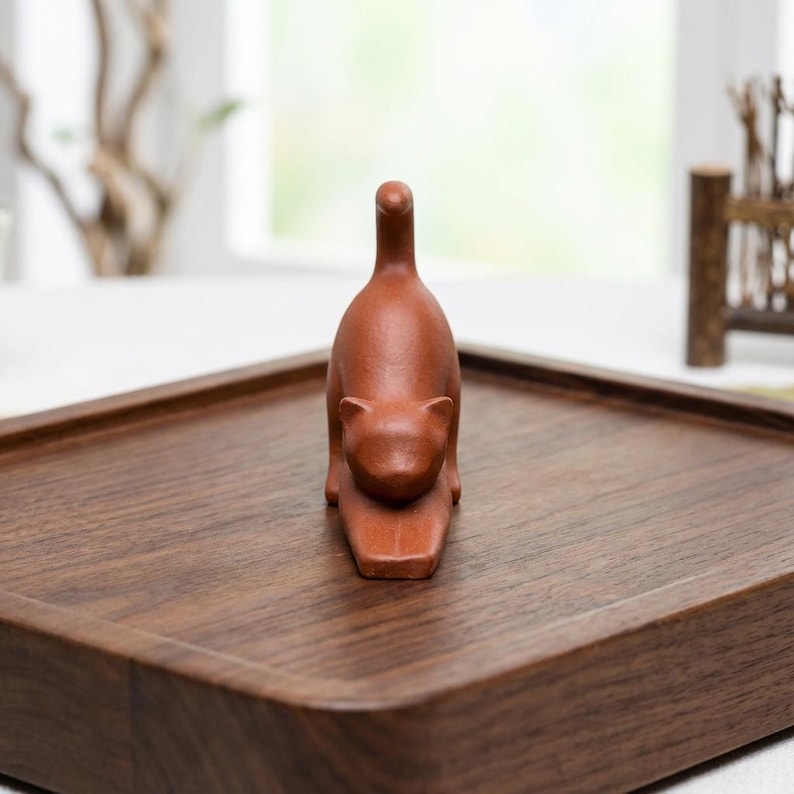 Puede incluir: Una peque&ntilde;a figura de gato marr&oacute;n rojizo en pose de estiramiento. El gato est&aacute; sobre una bandeja rectangular de madera. La bandeja tiene esquinas redondeadas y un acabado marr&oacute;n oscuro. El fondo est&aacute; borroso, con una ventana y algunos elementos decorativos visibles.