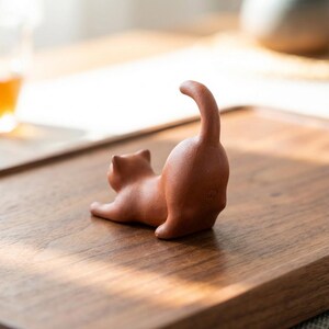 Puede incluir: Peque&ntilde;a figura de gato de terracota en una pose juguetona, con la cola arqueada hacia arriba. El gato est&aacute; tumbado sobre una superficie de madera. La figura es de color marr&oacute;n rojizo c&aacute;lido.