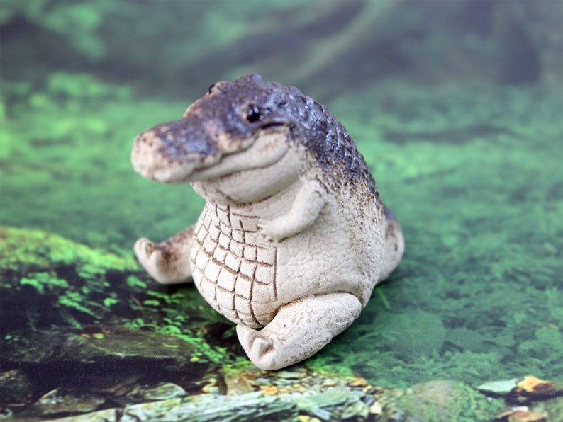 May include: A small, white ceramic alligator figurine with a gray and brown painted pattern. The alligator is sitting with its legs crossed and has a friendly expression.