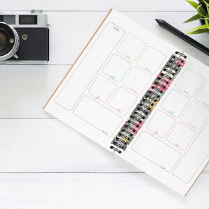 May include: A white planner with a floral patterned spine and a black pen on a white wooden surface. The planner has a weekly layout with boxes for each day of the week. The planner is open to the week of July 10th.