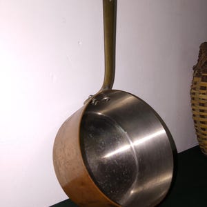 Può includere: Un pentolino rotondo in rame con interno argentato lucido e un manico lungo e sottile in ottone fissato con due rivetti. L'esterno del pentolino presenta una calda finitura metallica in rame, mentre l'interno mostra una superficie liscia.