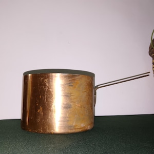 Peut inclure: Une casserole ronde en cuivre avec une finition métallique polie et une longue poignée fine en métal s'étendant sur le côté. La surface extérieure de la casserole présente des signes d'usure visibles, notamment des rayures et des nuances.
