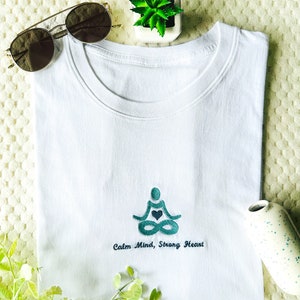May include: White t-shirt with an embroidered design of a person in a yoga pose with a heart in their chest. The text "Calm Mind, Strong Heart" is embroidered below the design.