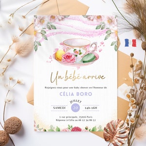 Peut inclure: Une invitation à une baby shower avec une illustration aquarelle d'une tasse à thé, des macarons et des fleurs. Le texte indique "Un bébé arrive" et comprend des détails sur l'événement, notamment la date, l'heure et le lieu.