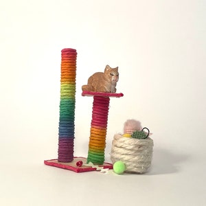 Könnte beinhalten: Ein Regenbogen-Katzenkratzbaum mit einer kleinen orangen Katzenfigur, die oben drauf sitzt. Ein Korb voller Katzenspielzeug steht vor dem Pfosten.