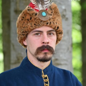 May include: A brown faux fur hat with a red top, featuring a central circular metal ornament with a turquoise stone and three brown and white striped feathers. The hat is worn with a dark blue coat with gold braided trim and metal buttons.