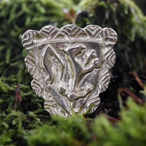 May include: A silver pendant in the shape of a shield with a stylized animal design. The pendant is sitting on a bed of green moss.