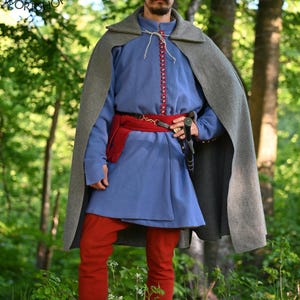 May include: A blue long-sleeved tunic with red trim and buttons, worn with red trousers, a wide red sash, and a grey wool cloak. A black cap with a feather and a metal brooch is worn on the head. A black belt with a metal buckle is at the waist.