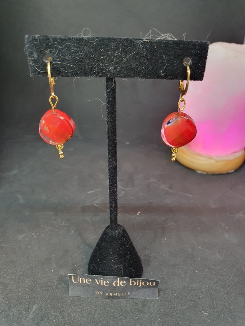 Peut inclure: Une paire de boucles d'oreilles cr&eacute;oles dor&eacute;es avec des perles de verre rouges et blanches. Les boucles d'oreilles sont pr&eacute;sent&eacute;es sur un support noir avec une pancarte indiquant "Une vie de bijou by Armelle".