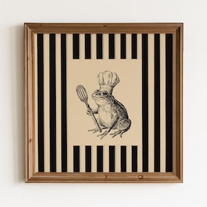 Può includere: Un'opera d'arte da parete quadrata incorniciata che presenta un'illustrazione a inchiostro nero di una rana che indossa un cappello da chef e tiene in mano una spatola. L'illustrazione centrale è su sfondo beige, circondata da un bordo a righe verticali nere e beige.