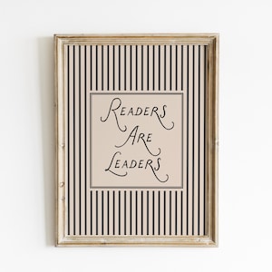 May include: A rectangular wooden frame containing a print with a beige background and vertical black stripes. In the center, a beige square box features the black text "Readers Are Leaders" written in a decorative, flowing script font.