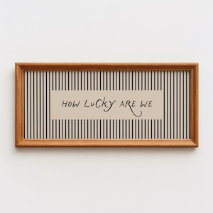 May include: A rectangular wooden frame containing a print with a vertical black and beige striped pattern. Centered on the print is a beige rectangular block featuring the black handwritten-style text that reads, How Lucky Are We.