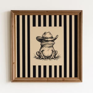 May include: A square framed wall art piece featuring a black and beige illustration of a frog wearing a cowboy hat. The central image is set against a background of vertical black and beige stripes, all enclosed within a medium-toned wooden frame.