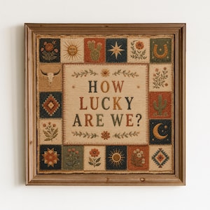 May include: A square framed textile wall hanging featuring the text HOW LUCKY ARE WE? in the centre, surrounded by a border of small squares with illustrations of flowers, cacti, a sun, a moon, a horseshoe, a skull, and geometric patterns.