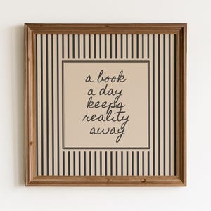 Könnte beinhalten: Ein quadratisches Wandbild in einem braunen Holzrahmen mit beigem Hintergrund und schwarzen vertikalen Streifen. In der Mitte befindet sich ein beiges Quadrat mit dem schwarzen kursiven Text: a book a day keeps reality away, umrandet von einer dünnen schwarzen Linie.