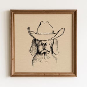 Könnte beinhalten: Ein quadratisches Wandbild mit einer schwarzen Tuschezeichnung eines Hundes, der einen Cowboyhut trägt. Die Illustration ist auf einem hellbeigen Hintergrund zentriert und befindet sich in einem schlichten, mittelbraunen Holzrahmen.