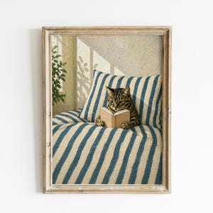 Könnte beinhalten: Gerahmter Kunstdruck, der eine getigerte Katze zeigt, die ein Buch auf einem gestreiften Kissen liest. Das Kunstwerk hat eine Vintage-Ästhetik mit weicher Beleuchtung und schafft eine gemütliche Atmosphäre, ideal für die Inneneinrichtung.