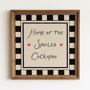 Könnte beinhalten: Ein quadratisches Wandschild in einem braunen Holzrahmen mit cremefarbener Mitte und dem schwarzen Text "Home of the Spoiled Cockapoo", flankiert von zwei kleinen rostfarbenen Sternen. Der Rand besteht aus einem schwarz-cremefarbenen Karomuster.