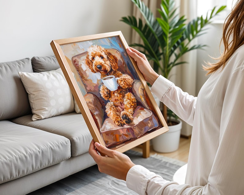 May include: A framed painting of a brown dog holding a white mug, seated in a brown armchair. The artwork has a light brown wooden frame. The painting's style is impressionistic, with visible brushstrokes and a warm colour palette.