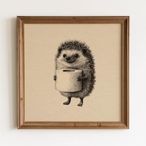 Può includere: Un'opera d'arte da parete quadrata incorniciata che presenta un'illustrazione a inchiostro in bianco e nero di un riccio in piedi che tiene un rotolo di carta igienica. L'opera è su uno sfondo beige chiaro in una cornice di legno marrone.