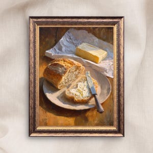 Könnte beinhalten: Ein gerahmtes Ölgemälde zeigt einen knusprigen Laib Brot, eine mit Butter bestrichene Brotscheibe und ein Stück gelbe Butter auf weißem Papier. Ein silbernes Buttermesser liegt auf dem Teller. Die Szene zeigt warmes, goldenes Licht und Texturen.