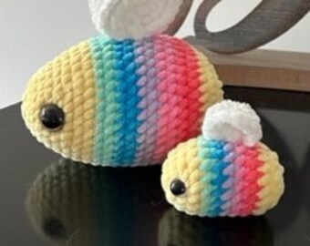 May include: Two crocheted rainbow bee plushies with white wings. The larger bee is laying down and the smaller bee is standing up.